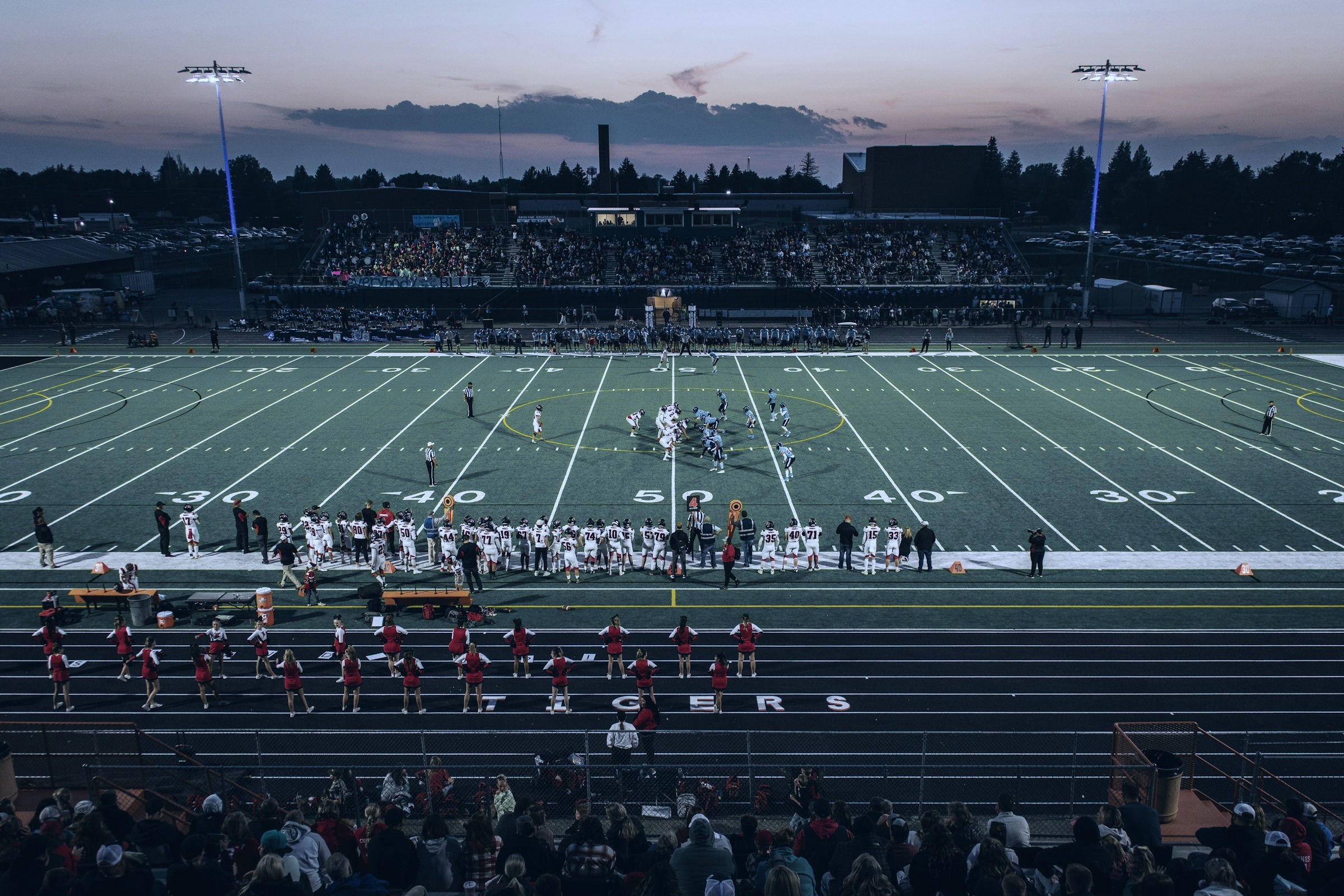 High school football under floodlights at dusk, seen from the home stands, with the packed crowd silhouetted across the foreground