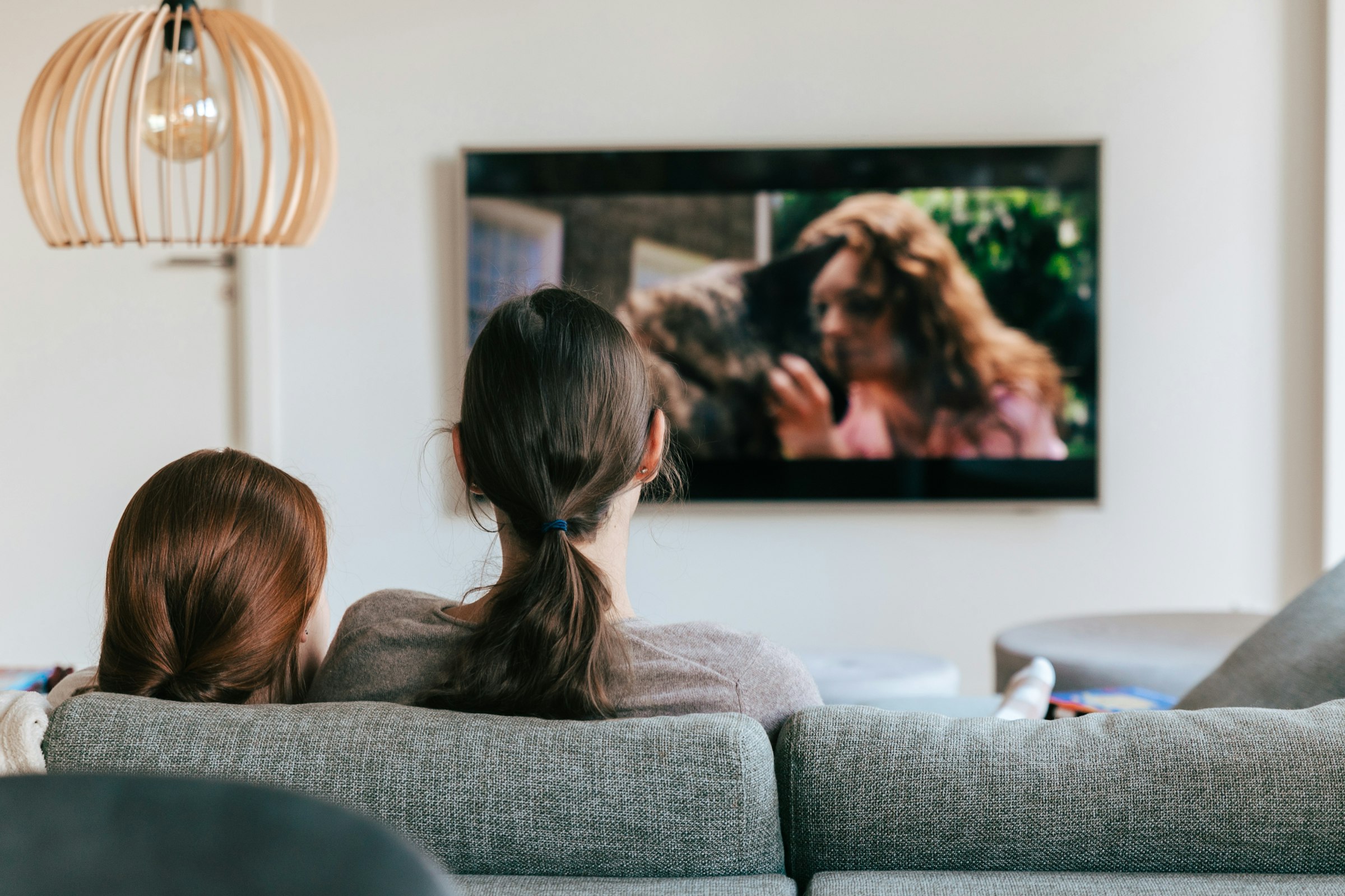 Two people on a couch watching a wall-mounted TV in a bright living room